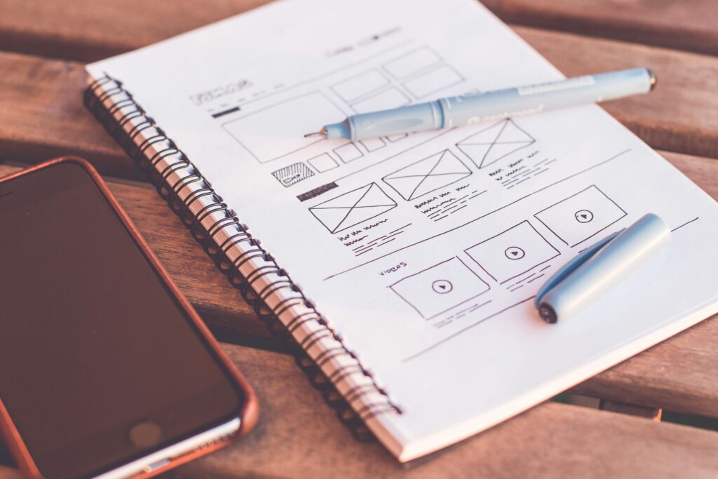 pexels-photo-196644-196644 Close-up of a notebook with wireframe sketches and a smartphone on a wooden desk.